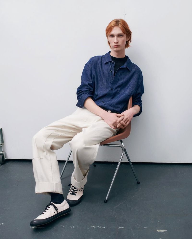 A young adult male with red hair sits on a chair in a studio, wearing a blue denim shirt, light-colored pants, and sneakers. 