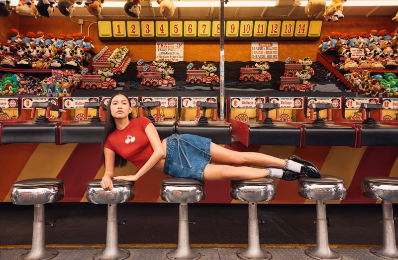 Asian woman in red top and denim skirt poses on chrome stools in front of a vintage carnival game with plush prizes in background.