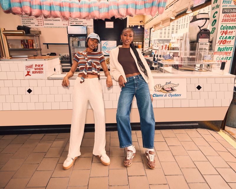 Two young Black women pose in front of a clam shack, wearing casual denim and cotton clothing; the background is filled with food signs.