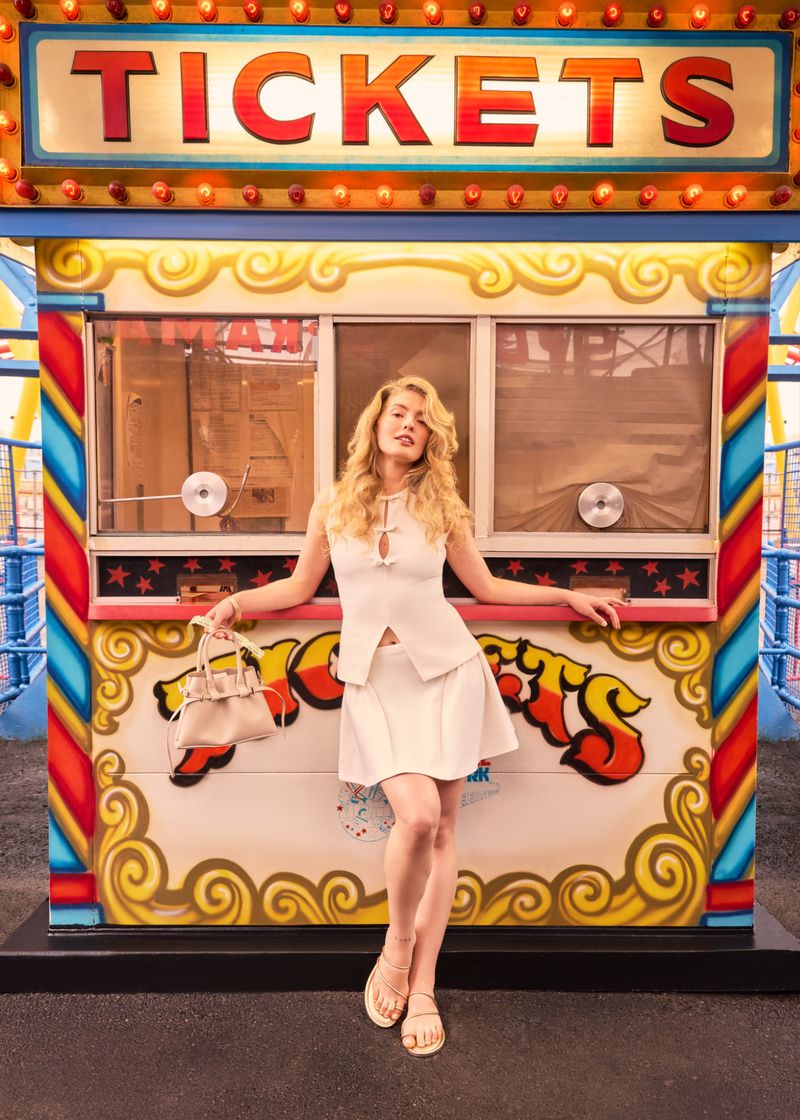 Woman in white outfit poses in front of a vibrant ticket booth with 'TICKETS' sign lit by marquee lights, holding a light colored handbag.