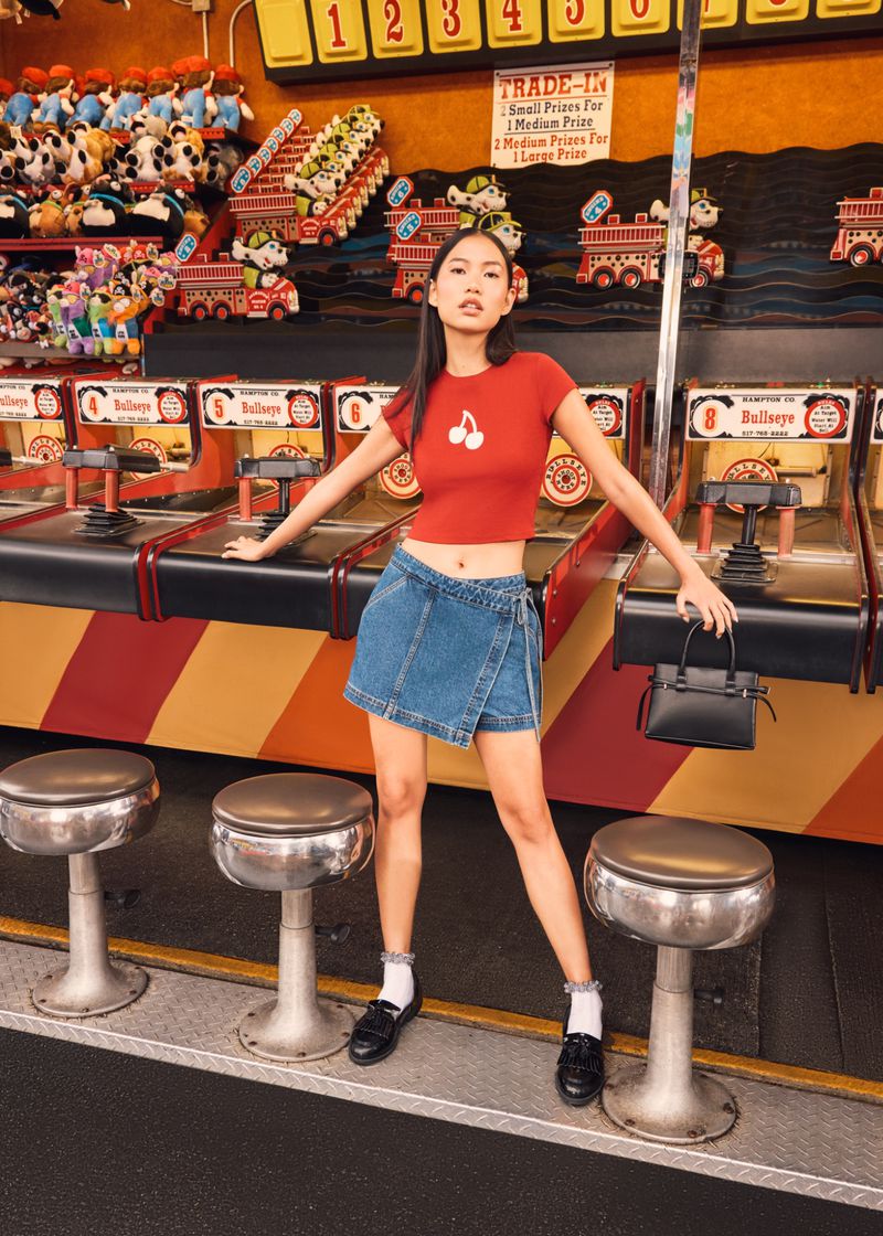 Young woman in denim skirt and red cherry t-shirt stands at a vintage boardwalk game with stuffed animal prizes in the background.