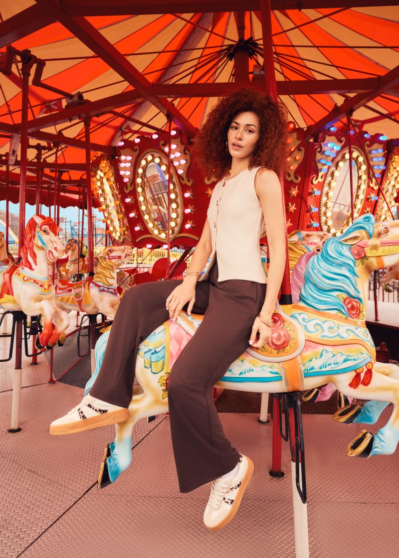 A young woman with curly hair sits on a carousel horse wearing a cream top and brown pants, set against a red and white carousel.