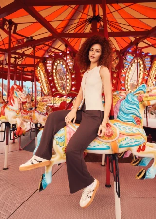 A young woman with curly hair sits on a carousel horse wearing a cream top and brown pants, set against a red and white carousel.