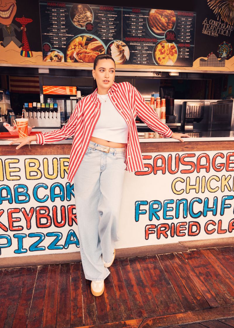Woman in a red-and-white striped shirt and light blue jeans standing in front of a vintage food stand with a hand-painted menu.