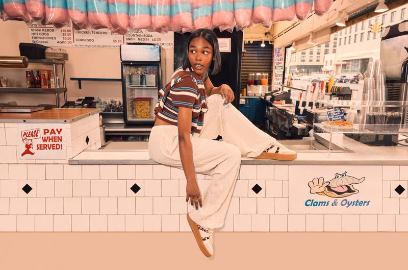 Young woman in striped crop top and off-white pants poses on a counter in front of a clams and oysters stand inside an eatery.