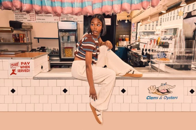 Young woman in striped crop top and off-white pants poses on a counter in front of a clams and oysters stand inside an eatery.