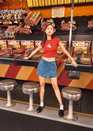 Model wearing a red cropped tee and denim mini skirt holding a black handbag at a retro carnival game counter.