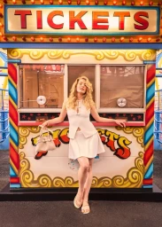 Blonde woman in a white skirt set and sandals posing with a handbag in front of a colorful carnival ticket booth.