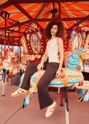 Woman with curly hair in a sleeveless cream vest and dark brown trousers sitting on a colorful vintage carousel horse.