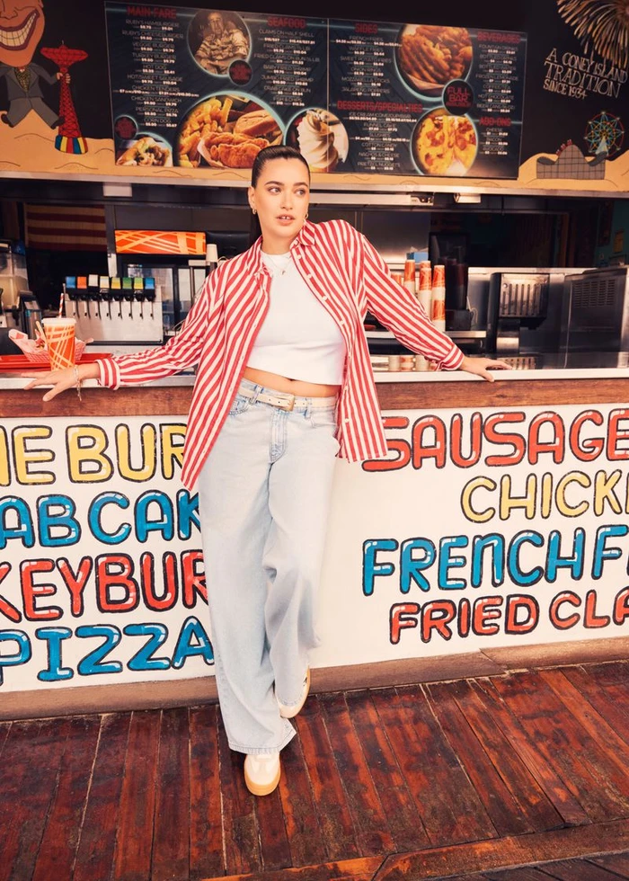 Woman in a red and white striped shirt and baggy light wash jeans leaning against a retro food stand counter on a wooden boardwalk.
