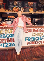 Woman in a red and white striped shirt and baggy light wash jeans leaning against a retro food stand counter on a wooden boardwalk.