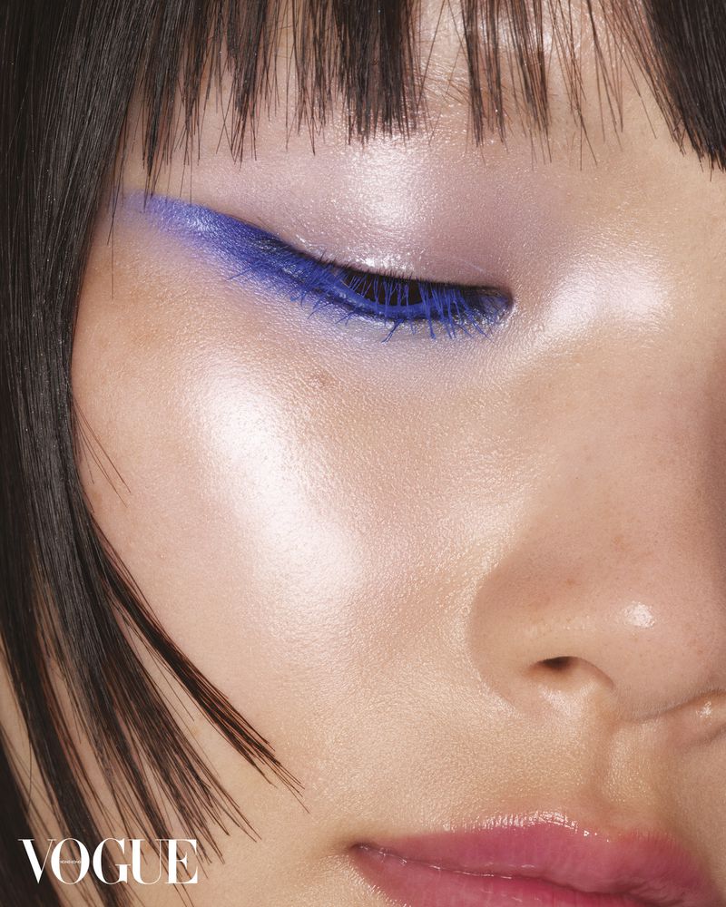 Close-up shot of an adult woman's face with bold blue eye makeup, glossy lips, and dark bangs, featured in Vogue magazine.