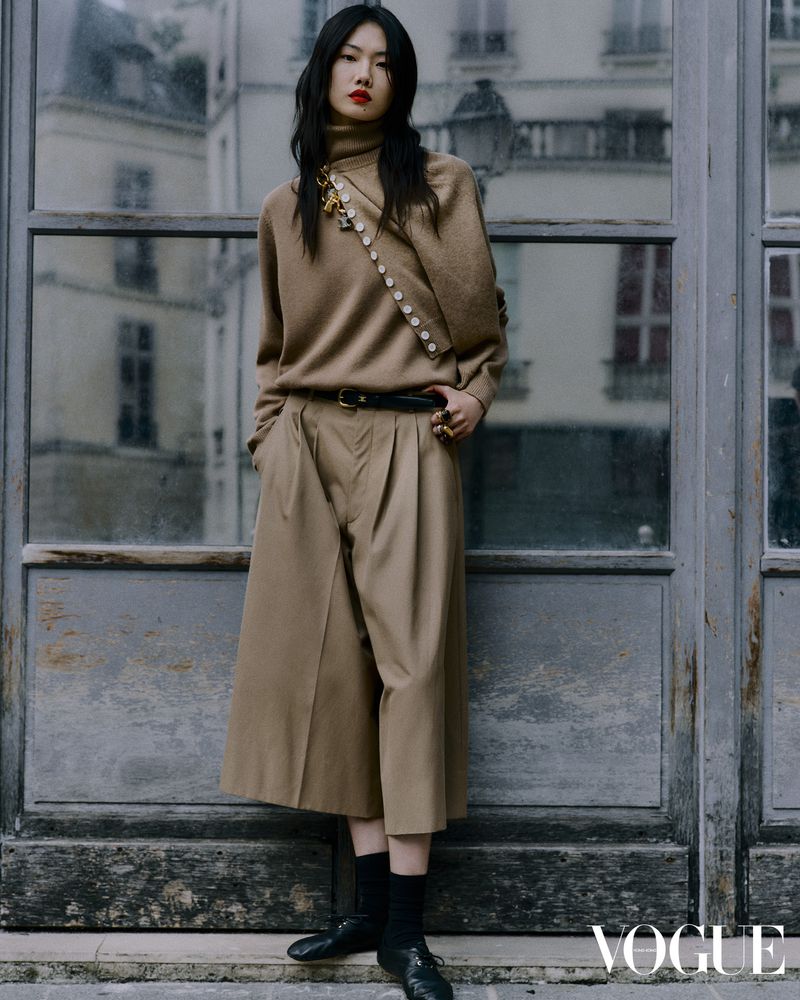 Fashion editorial shot of a young Asian woman in a beige sweater and wide-legged pants, posing in front of a weathered glass door.