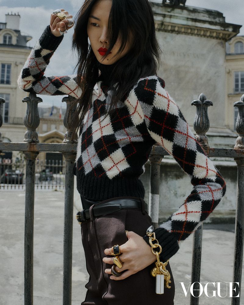 Fashion editorial photo of an Asian female model wearing an argyle sweater, brown pants, and rings, posing in an urban outdoor setting.