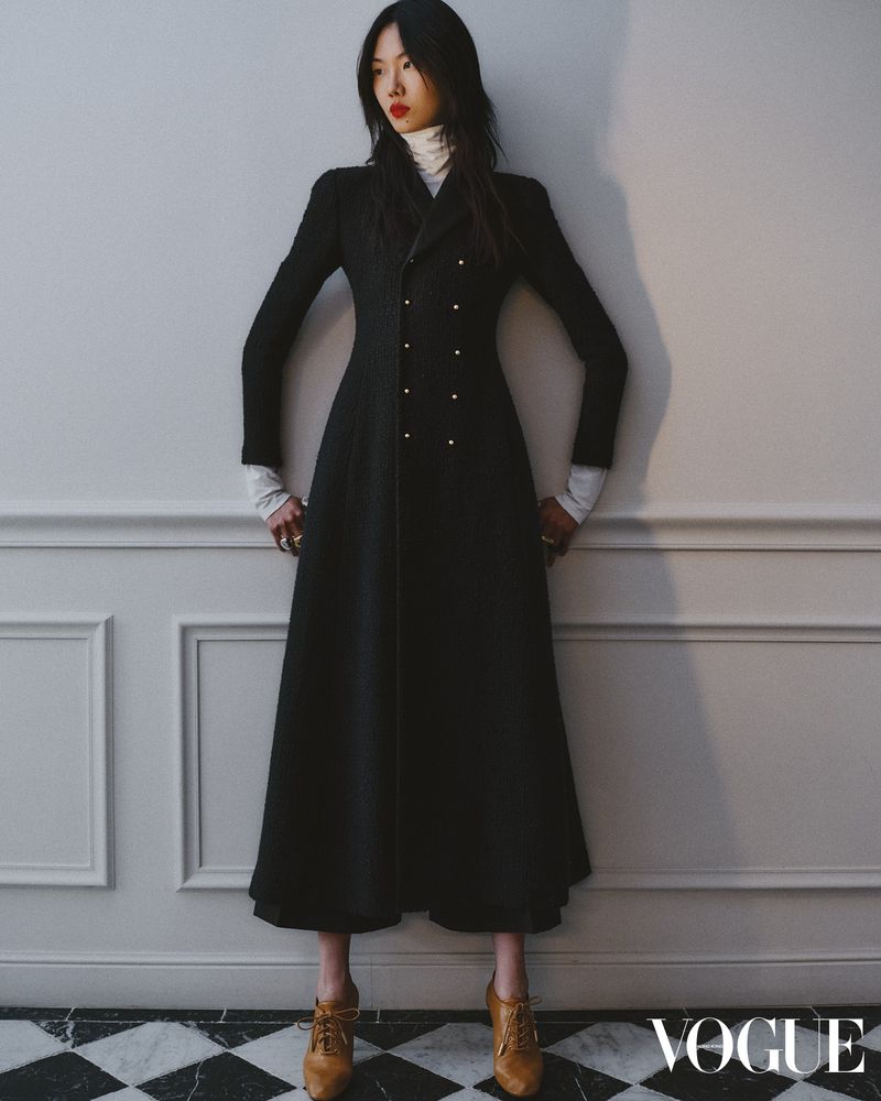 A woman stands in a long black coat and tan lace-up shoes in a studio with a marbled floor and white wall, posing for a fashion editorial.