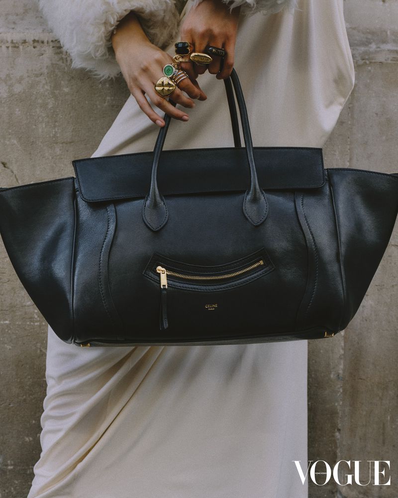 Black leather tote bag held by a person with rings, posing against a textured grey wall, with a draped white dress.