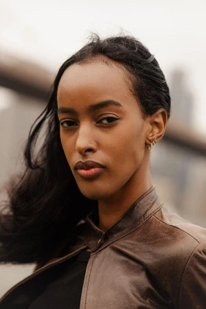 Portrait of a young African woman with dark hair wearing a leather jacket, posing outdoors in front of a blurred cityscape on a cloudy day.