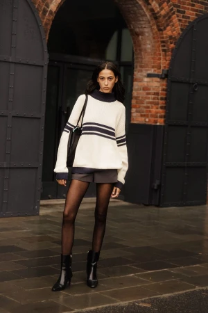 A young woman models a striped sweater with shorts, tights, and ankle boots in front of a brick building and metal gates outdoors.