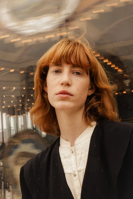 Close-up portrait of a fair-skinned adult woman with red hair wearing a black blazer and cream-colored shirt, captured indoors with soft lighting.