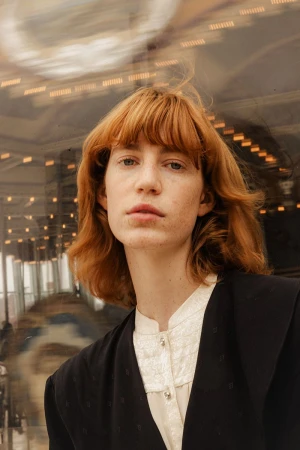 Close-up portrait of a fair-skinned adult woman with red hair wearing a black blazer and cream-colored shirt, captured indoors with soft lighting.