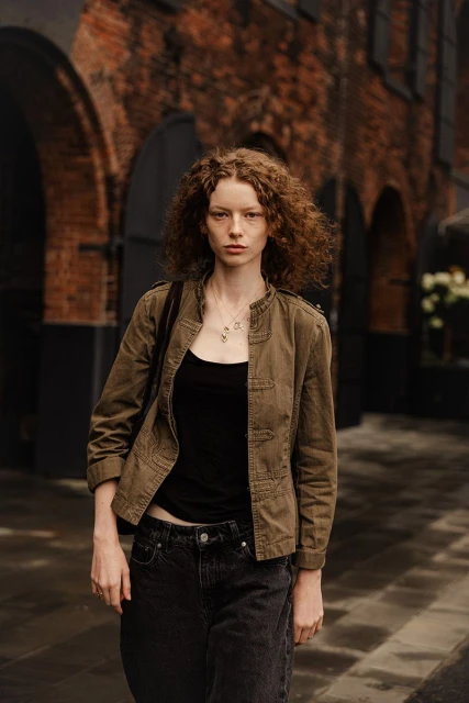 Adult woman with auburn curly hair poses in a black tank top and olive jacket in front of a brick building with arched doorways.