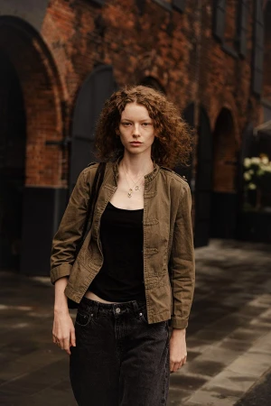 Adult woman with auburn curly hair poses in a black tank top and olive jacket in front of a brick building with arched doorways.