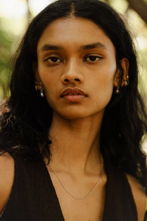 Close-up portrait of an adult woman with dark hair and brown eyes, wearing a brown tank top, posing outdoors with green foliage in the background.