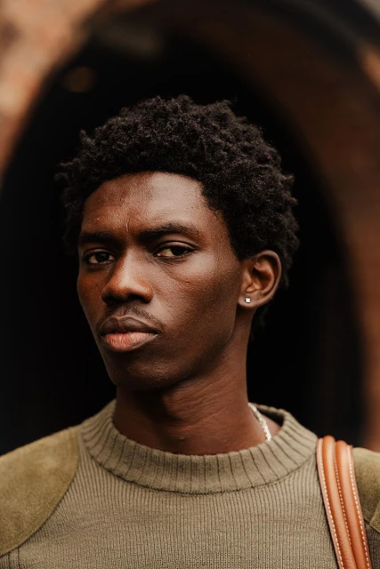 Close-up portrait of a Black man with dark hair wearing an olive sweater with suede shoulder patches, standing in front of a dark background.
