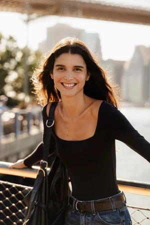 Smiling woman with long dark hair leans against a rail, wearing a black long-sleeve shirt, jeans, with the Brooklyn Bridge visible in the background.