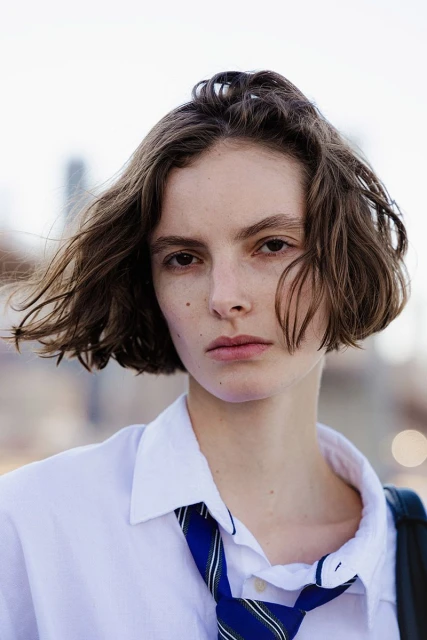 Portrait of a young adult female with wavy brown hair, wearing a white shirt and blue tie, against a blurred urban background.