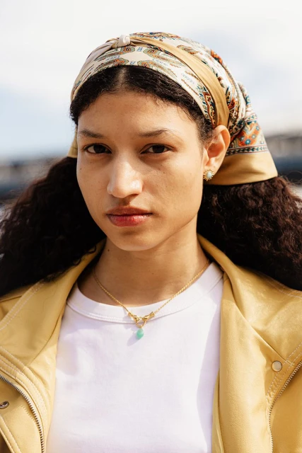 Close-up portrait of a woman with curly hair wearing a patterned headscarf, gold necklace, and yellow jacket against a sunny sky.