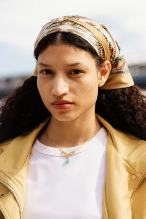 Close-up portrait of a woman with curly hair wearing a patterned headscarf, gold necklace, and yellow jacket against a sunny sky.