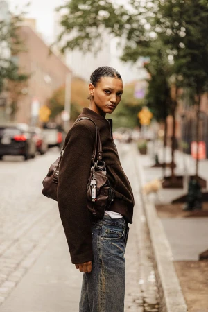 Woman in a brown jacket and blue jeans stands on a city street with trees in the background and a brown purse on her shoulder.