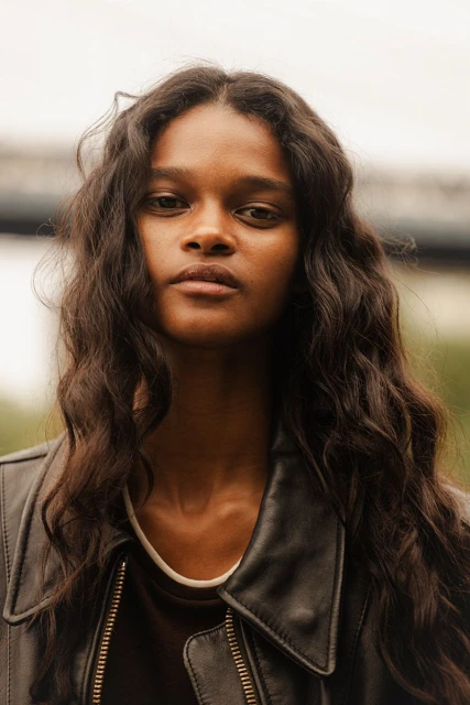 Portrait of a woman with long wavy hair, wearing a leather jacket, looking directly at the camera in an outdoor setting.