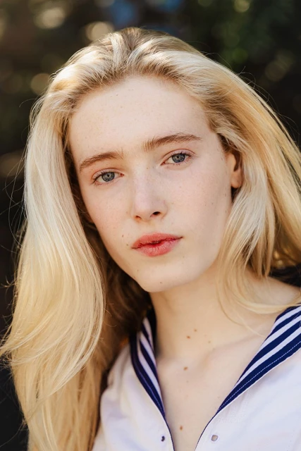 Close-up portrait of a fair-skinned woman with long blonde hair, wearing a white shirt with a blue-striped collar, outdoors.