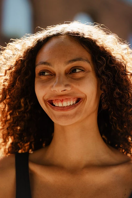 Portrait of a smiling woman with curly hair, backlit by sunlight, creating a bright and cheerful vibe. The background is blurred.