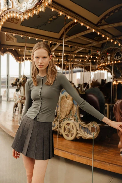 Fashionable young woman poses near vintage carousel in long-sleeve top and pleated skirt. Indoor setting, neutral color palette.