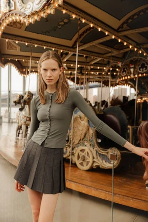 Fashionable young woman poses near vintage carousel in long-sleeve top and pleated skirt. Indoor setting, neutral color palette.