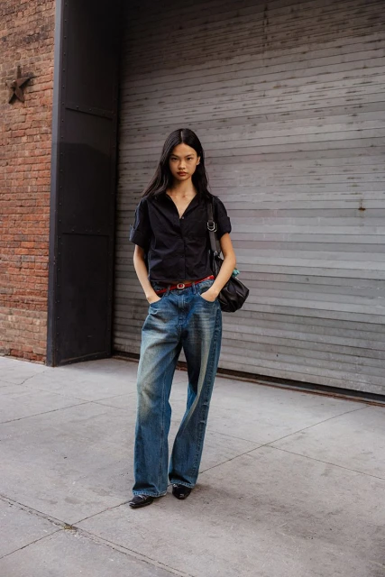 A young woman in wide-leg jeans and a dark shirt stands against a grey brick wall with a red brick building visible to the left.