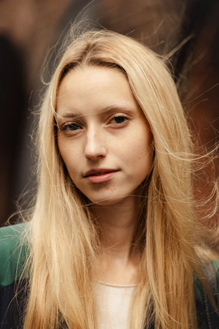 Close-up portrait of a blonde woman with fair skin wearing a neutral top and jacket, set against a blurred brown background.