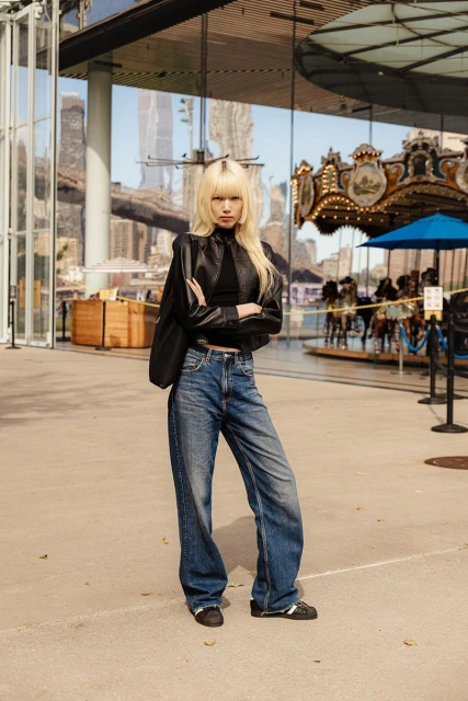 Blonde woman stands in denim and black leather jacket in front of a carousel in an urban setting on a sunny day.