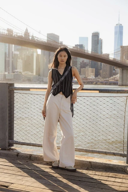 Woman in vest and wide-leg jeans poses on a pier with the Brooklyn Bridge and New York City skyline in the background on a sunny day.