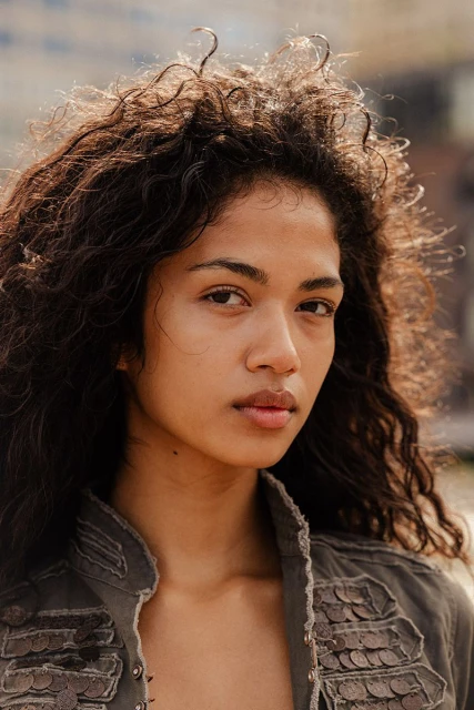 Close-up portrait of an adult female with curly brown hair, wearing a green jacket, posing outdoors in natural light against a blurred backdrop.