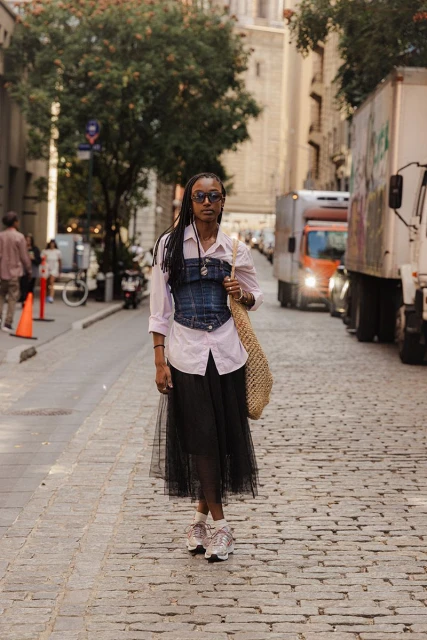 A woman in a light pink button-down shirt, denim corset, and black tulle skirt walks down a city street with tall buildings.