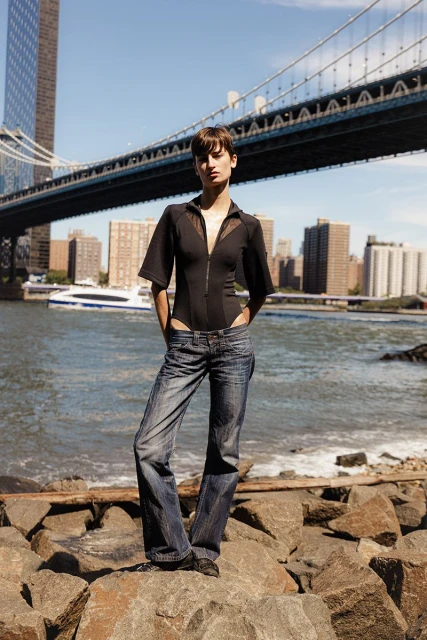Adult woman in a black bodysuit and jeans standing on rocks with a bridge and city buildings in the background, clear sky.
