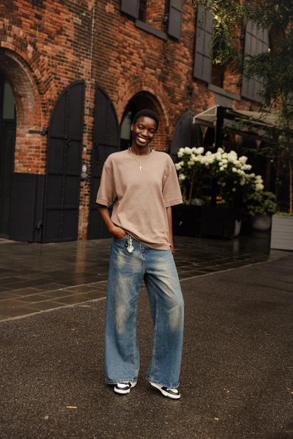 Full-length portrait of a Black woman outdoors in casual clothes, posing in front of a brick building with greenery; fashion editorial.