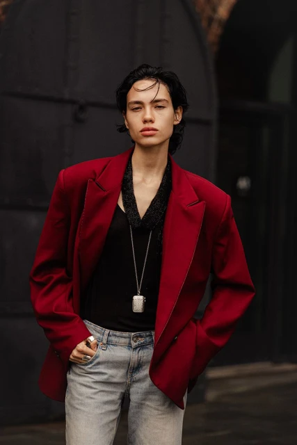 A person in a red jacket and light-wash jeans stands in front of a black metal door. A silver necklace and scarf complete the outfit.