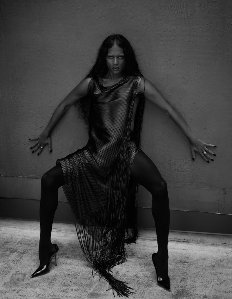 Black and white shot of a woman with dark hair wearing a fringe dress and heels, leaning against a grey wall in a fashion pose.