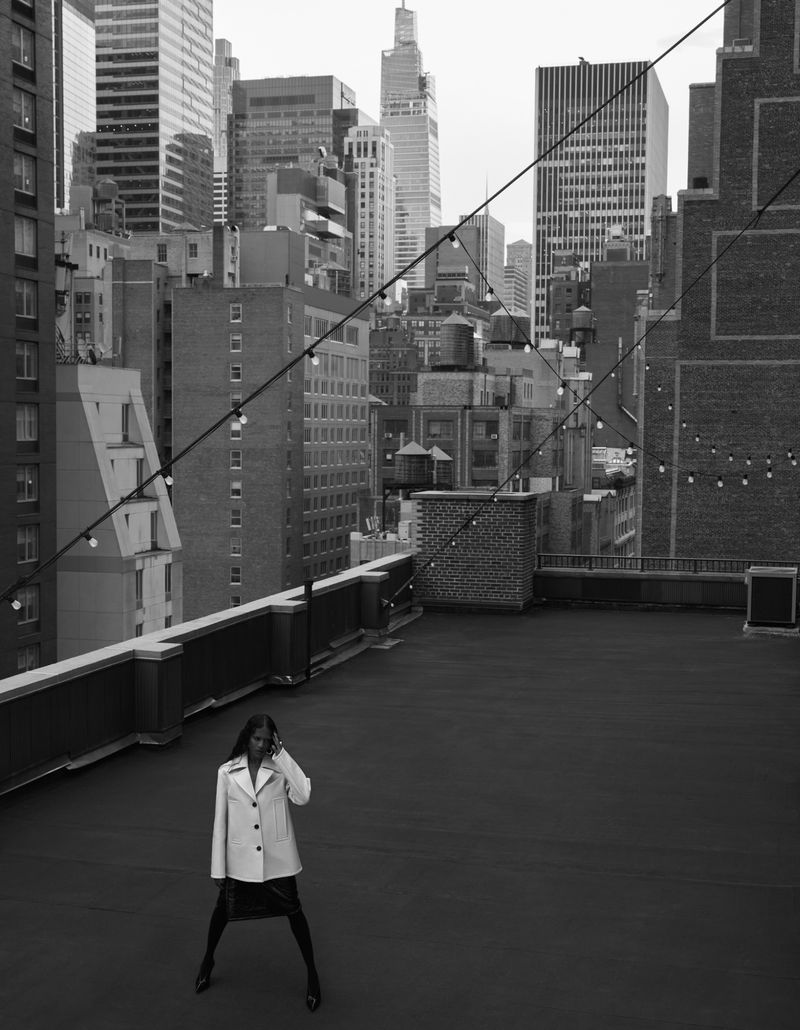 A woman in a trench coat stands on a rooftop overlooking a city skyline in a black and white photograph, talking on a cell phone.