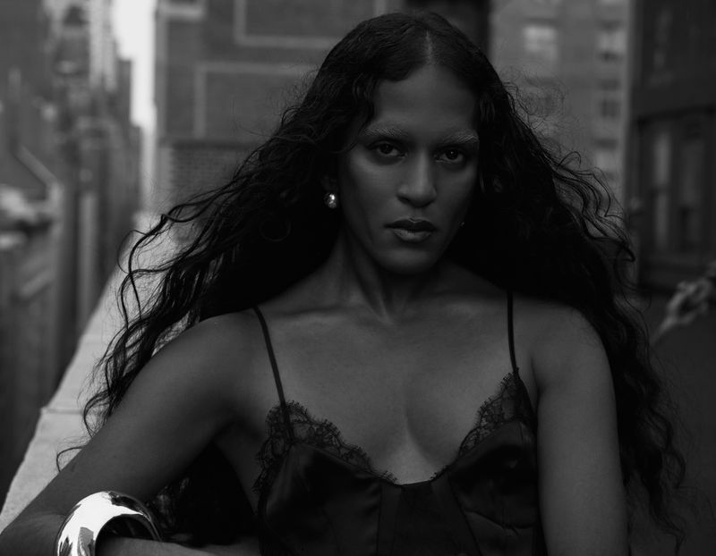 Black and white portrait of an adult female wearing a silk top and silver bracelet, posing outdoors with an urban backdrop.